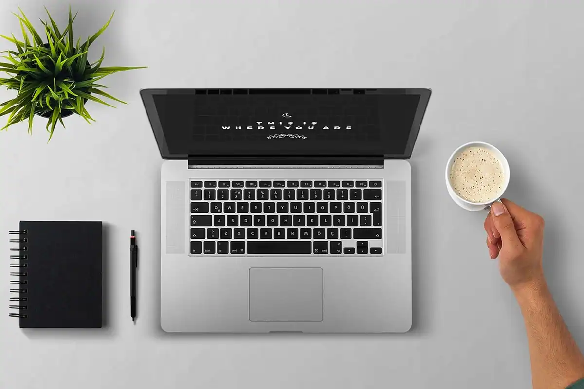 Laptop deschis, carnet negru și o ceașcă de cafea, ținută de o mână, pe un birou