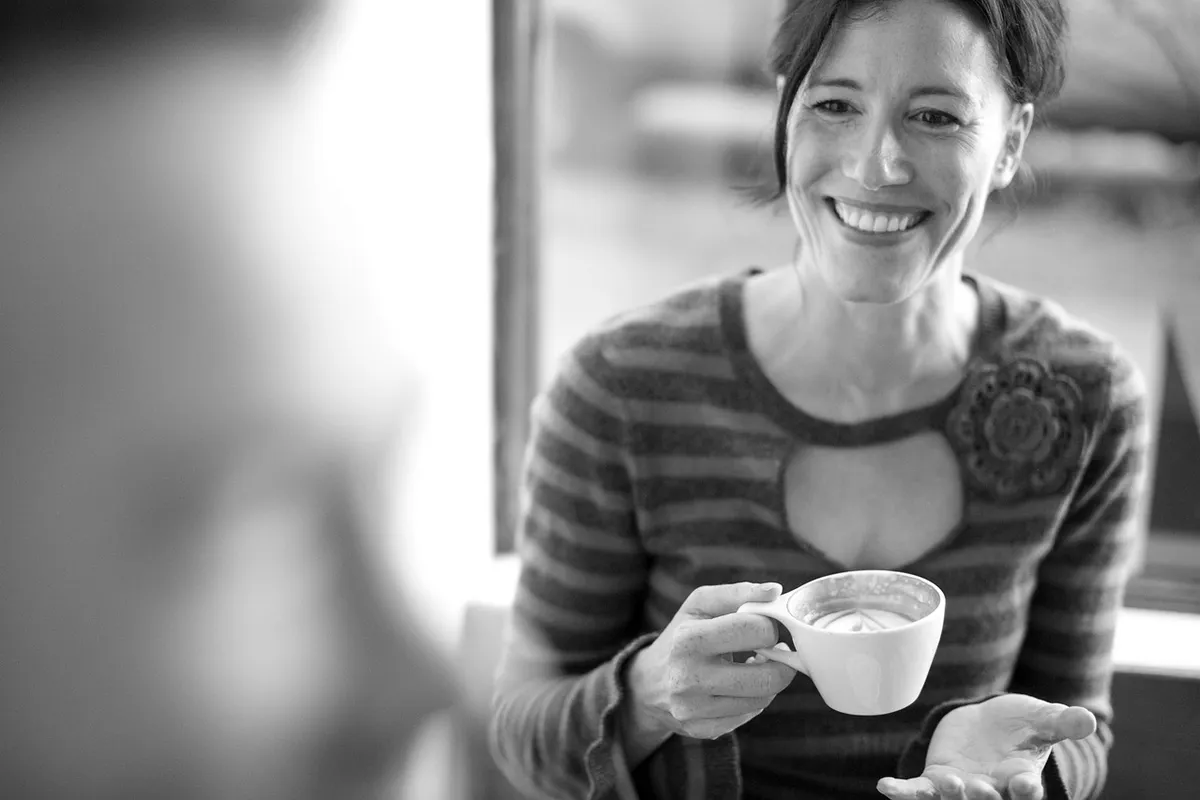 Femeie zâmbitoare ține o ceașcă de cafea, într-o fotografie alb-negru, la o discuție relaxată