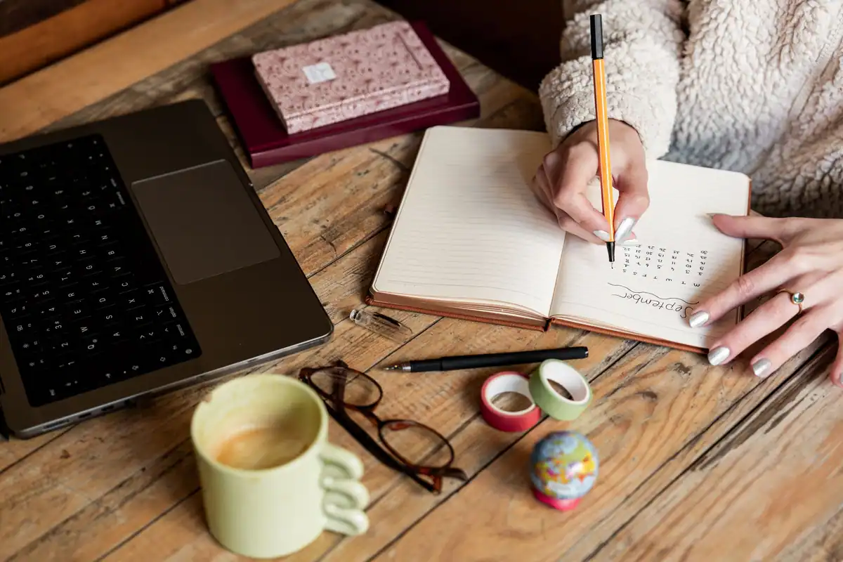 Femeie care notează planul unei călătorii într-un carnet, lângă laptop și o cană de cafea