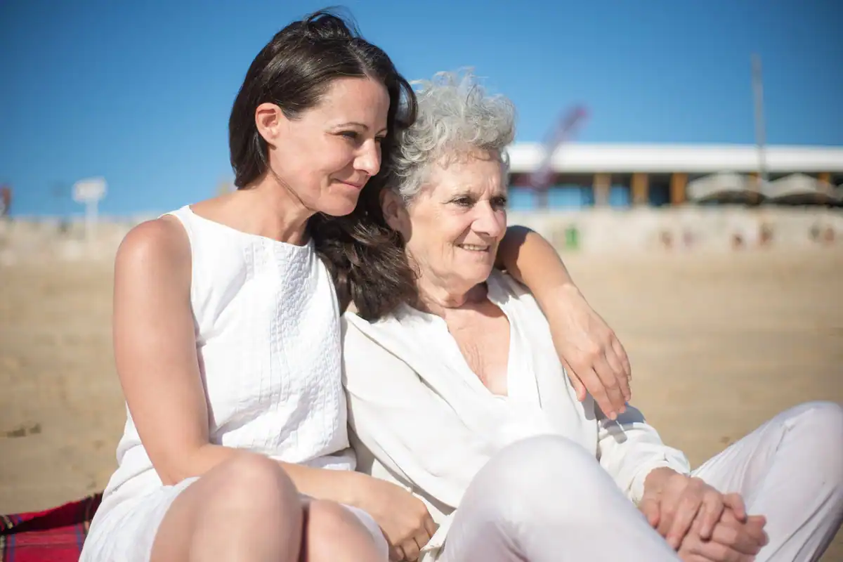 Femeie adultă și mama ei în vârstă stau îmbrățișate pe o plajă, zâmbind