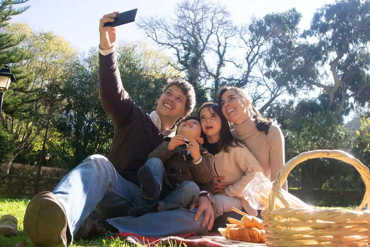 Familie care își face un selfie pe iarbă, la picnic, lângă un coș de răchită