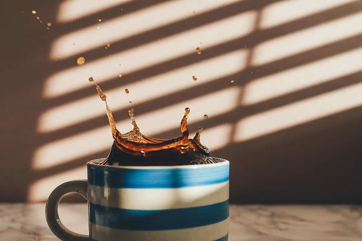 Ceașcă dungată din care țâșnesc stropi de cafea, pe o masă luminată de soarele dimineții