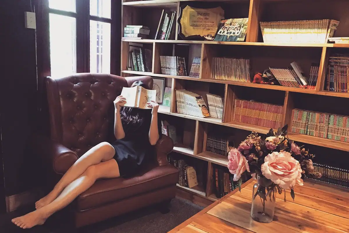 Femeie care citește întinsă într-un fotoliu de piele, lângă o bibliotecă și un buchet de flori